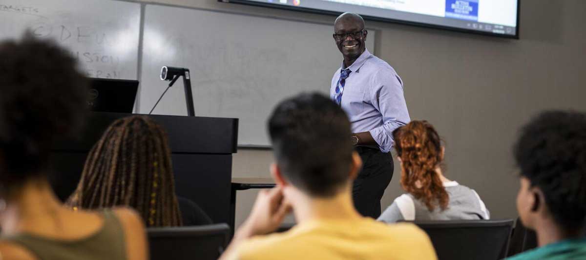 Smiling professor, standing, looks at seated students in classroom
